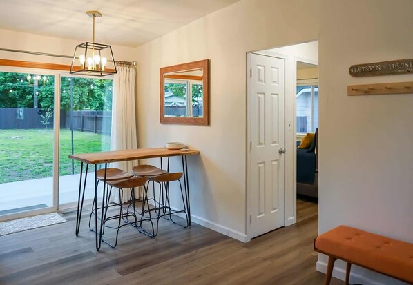 A room with a wooden dining table and bar stools, a large window with curtains, a door leading to another room, and a small bench.