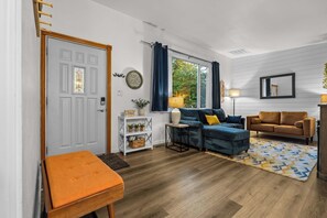 A living room with a white door, a blue sofa, a brown sofa, a small orange bench, and a patterned rug.