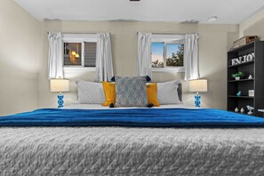 A bedroom with a bed, two windows with curtains, a nightstand with a lamp, and a bookshelf with decorative items.
