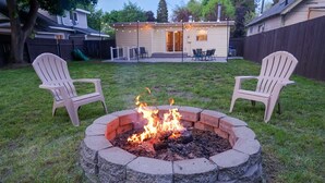 Patio trasero con un hogar a leña, dos sillas Adirondack y una casa al fondo.
