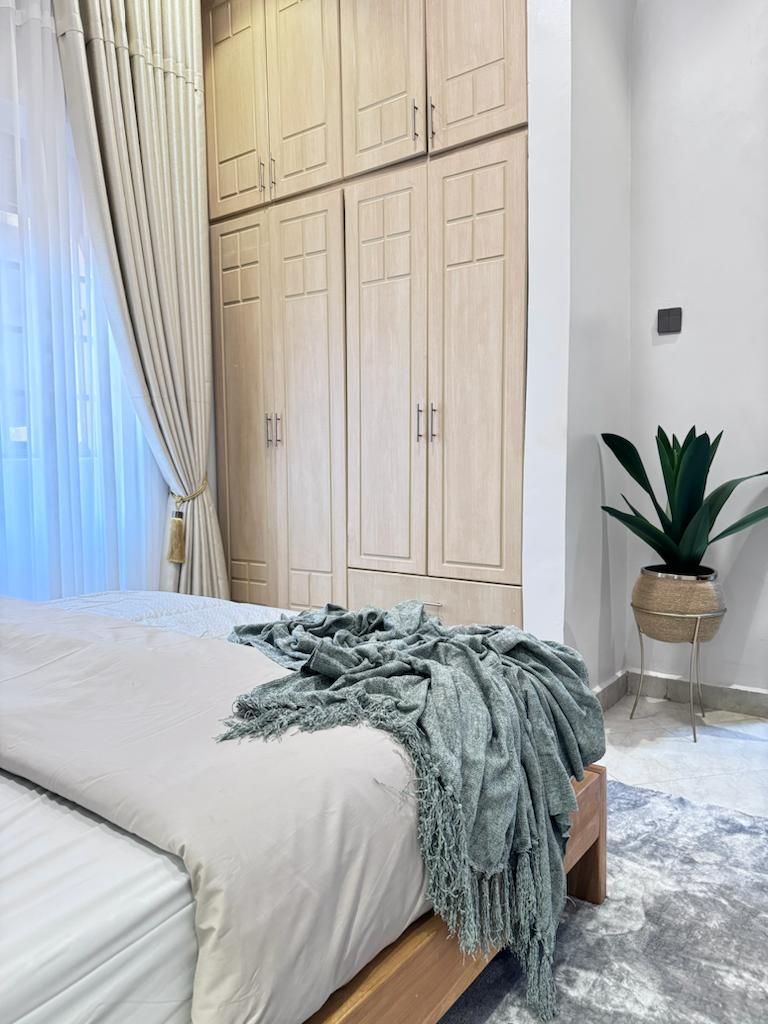 A bedroom with a bed, a large wardrobe, a potted plant, and a window with curtains.