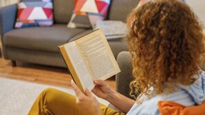 Una persona sentada en un sofá leyendo un libro en una sala de estar con un sofá gris y almohadones coloridos.
