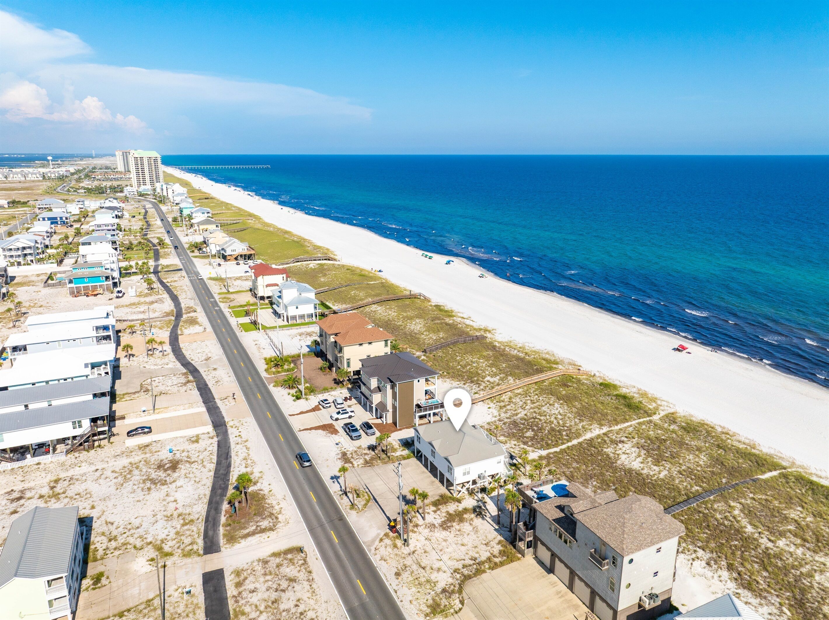 An aerial view of a coastal area with a long stretch of beach, a road running parallel to the shore, and several buildings including a large hotel with a pool.