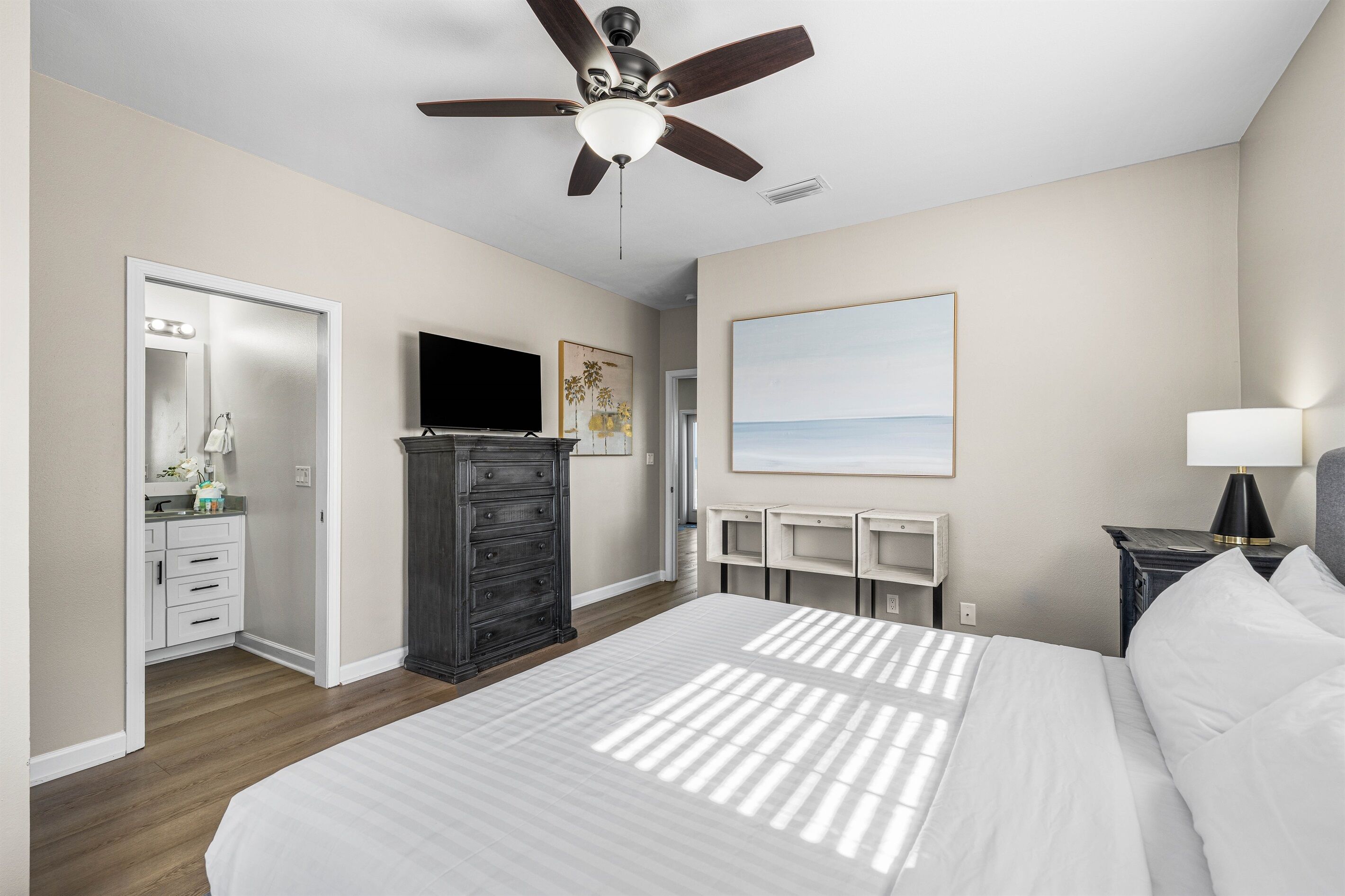 A bedroom with a bed, a ceiling fan, a dresser, a TV, a painting, and a nightstand.