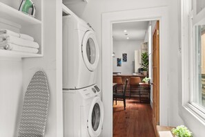 A laundry room with two washing machines, a surfboard, and a dining area with wooden flooring and chairs.