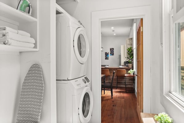 A laundry room with two washing machines, a surfboard, and a dining area with wooden flooring and chairs.