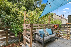 A wooden deck with a sofa and cushions, surrounded by a wooden fence and greenery.