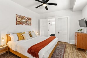 Een slaapkamer met een bed, een plafondventilator, een schilderij, een televisie op een houten dressoir en een deur die naar een andere kamer leidt.