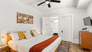 Une chambre à coucher avec un lit, un ventilateur de plafond, un tableau, un téléviseur posé sur une commode en bois et une porte donnant sur une autre pièce.