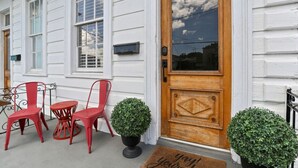 Un bâtiment blanc avec une porte d’entrée en bois, deux chaises rouges et deux plantes en pot.