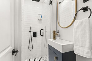 Een moderne badkamer met witte betegelde wanden, een witte wastafel met gouden kraan, een handdoek aan een handdoekhouder en een douche met een handdouche.