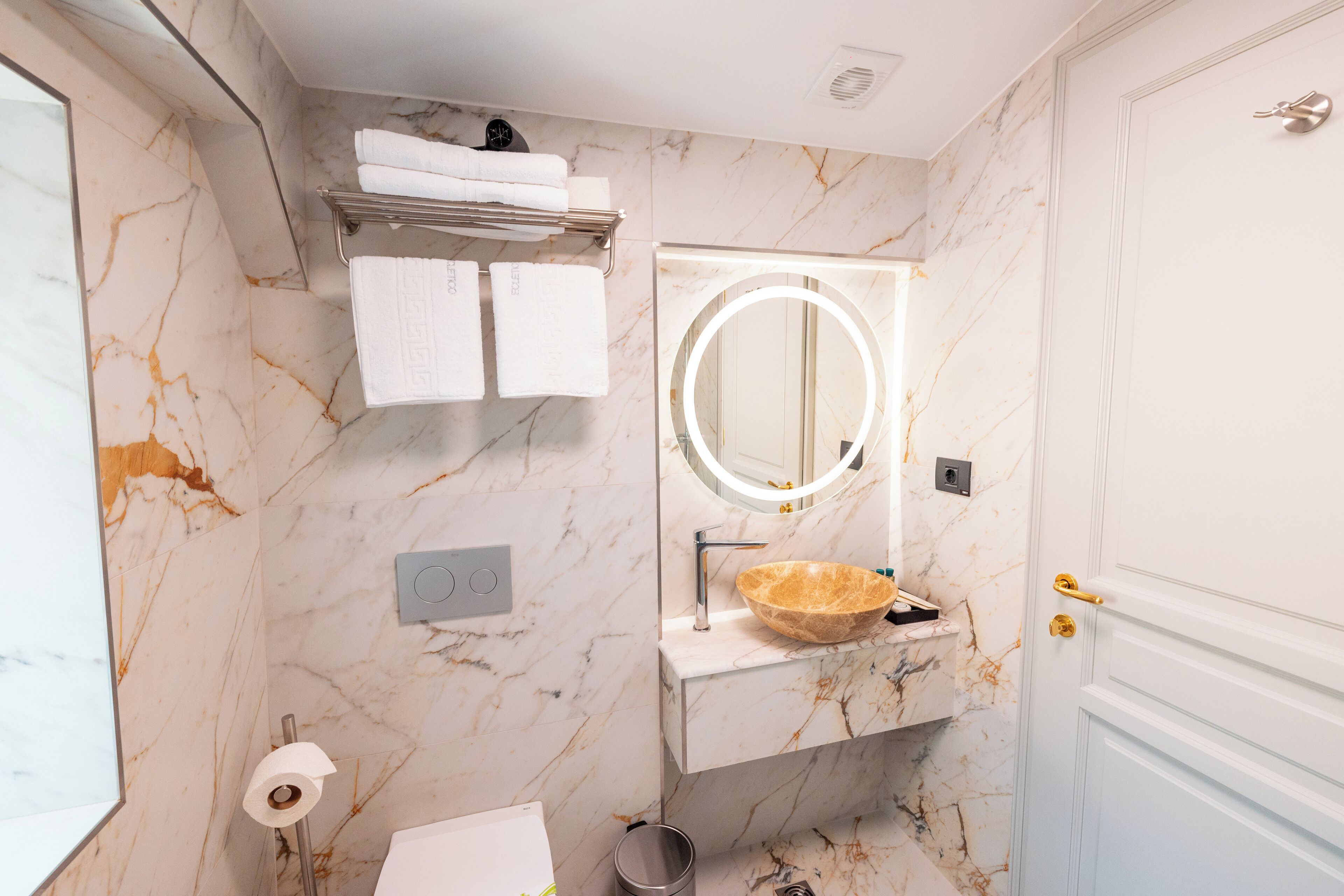 Une salle de bain avec des murs en marbre, un lavabo avec un receveur, un miroir avec un éclairage intégré et un porte-serviettes.