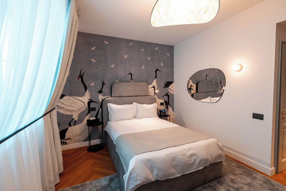 A bedroom with a bed, a bedside table, a lamp, a mirror, and a wall with a bird mural.