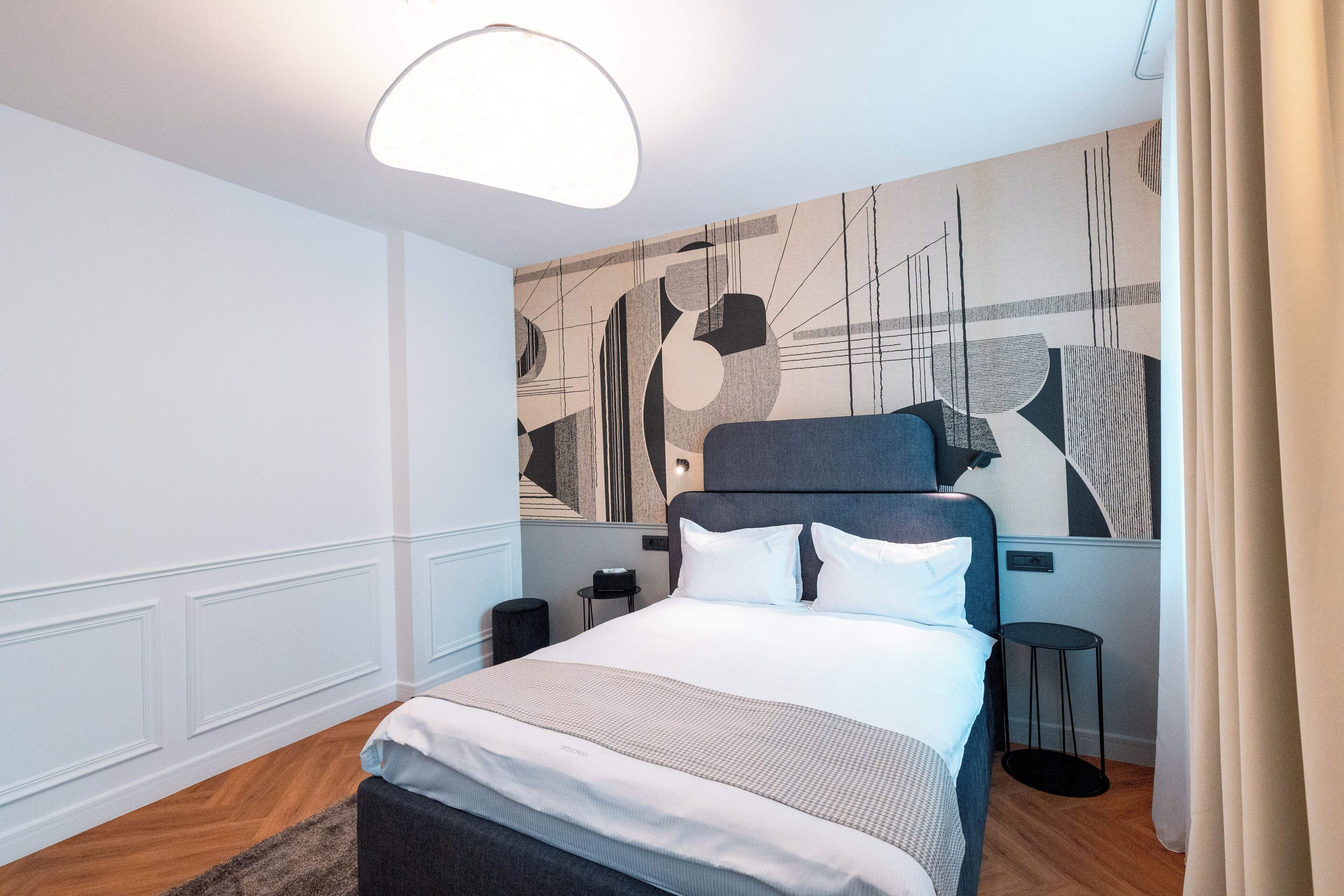 Ein modernes Hotelzimmer mit einem großen Bett, einem Nachttisch, einem kleinen Hocker und einer Wand mit abstrakter Kunst.