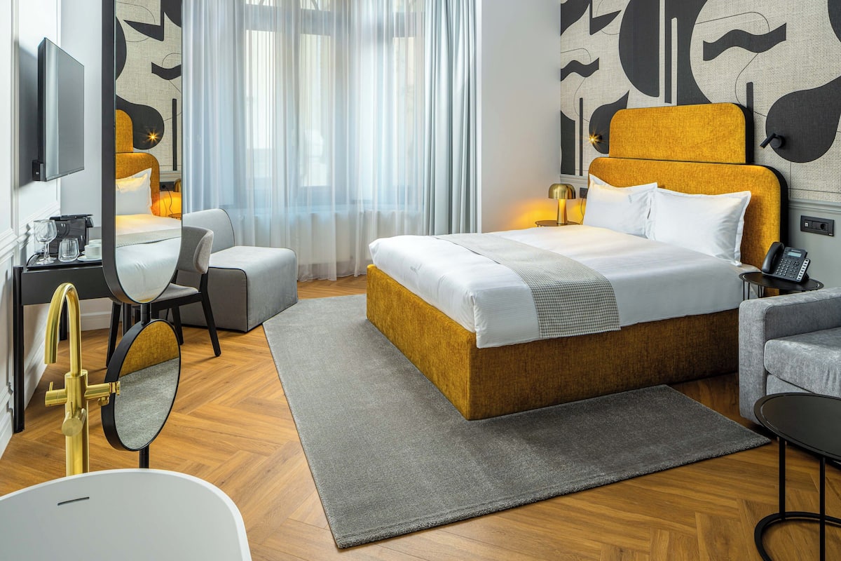 A modern hotel room with a large bed, a desk with a chair, a sofa, a TV mounted on the wall, and a bathroom with a golden faucet.