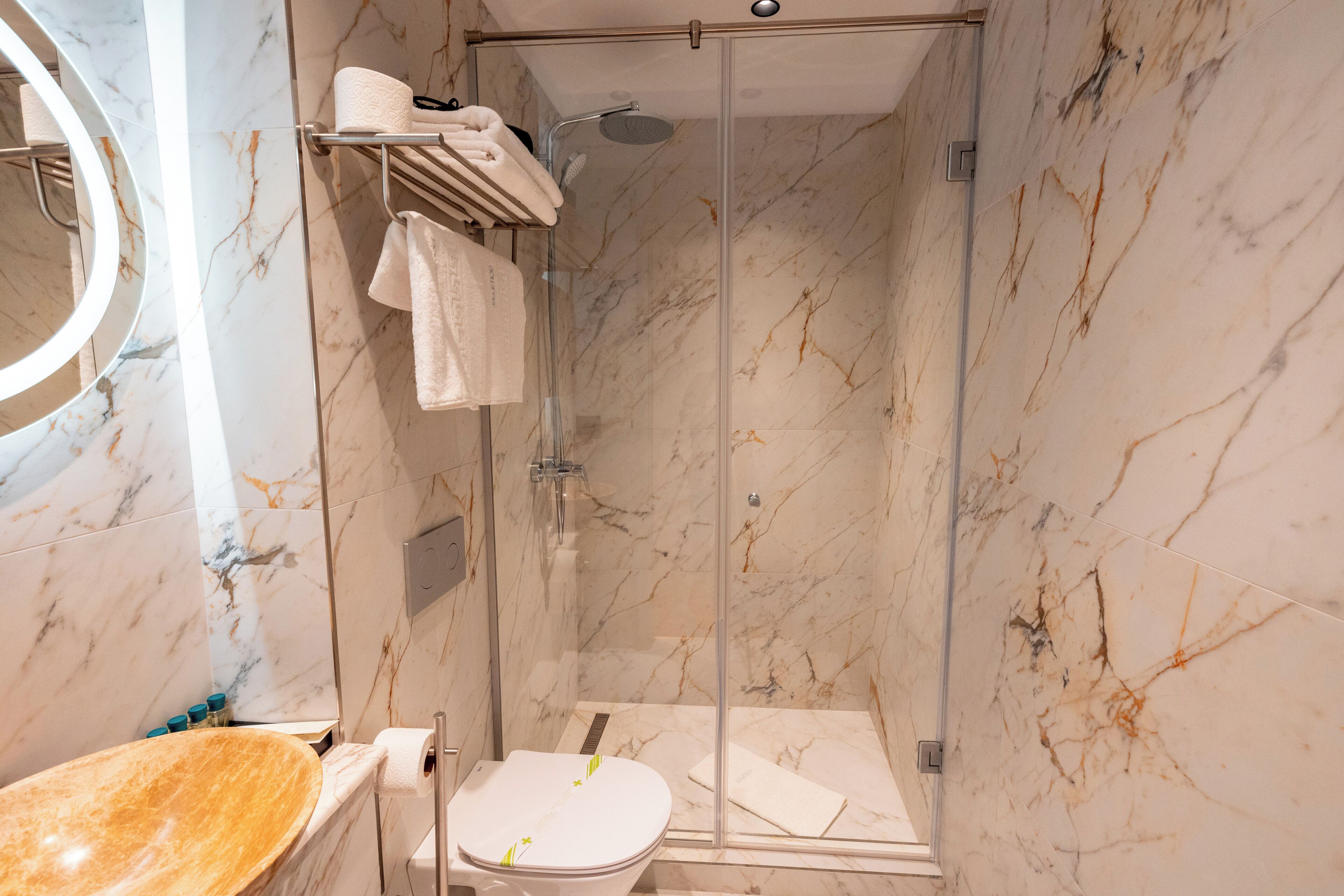 A modern bathroom with a glass-enclosed shower, a marble-tiled wall, a wooden vanity, and a round mirror.