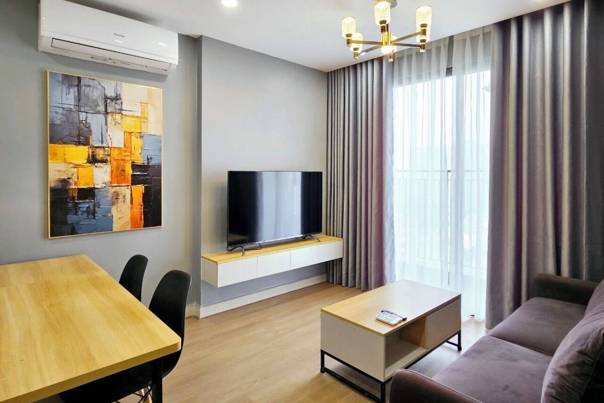 A modern living room with a flat-screen TV on a built-in shelf, a dining table, a sofa, and a large window with curtains.