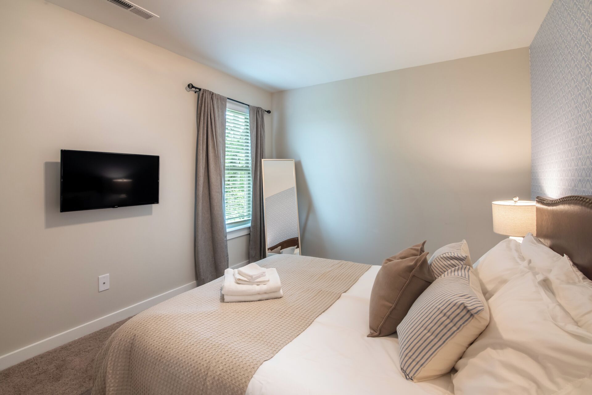 Une chambre d’hôtel avec un lit, un téléviseur à écran plat fixé au mur, une table de chevet avec une lampe et une fenêtre avec des rideaux.