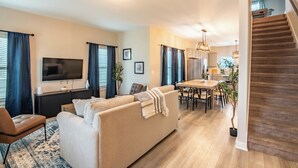 Un salon moderne avec un canapé, un téléviseur et un coin repas. Un escalier mène à l’étage supérieur.