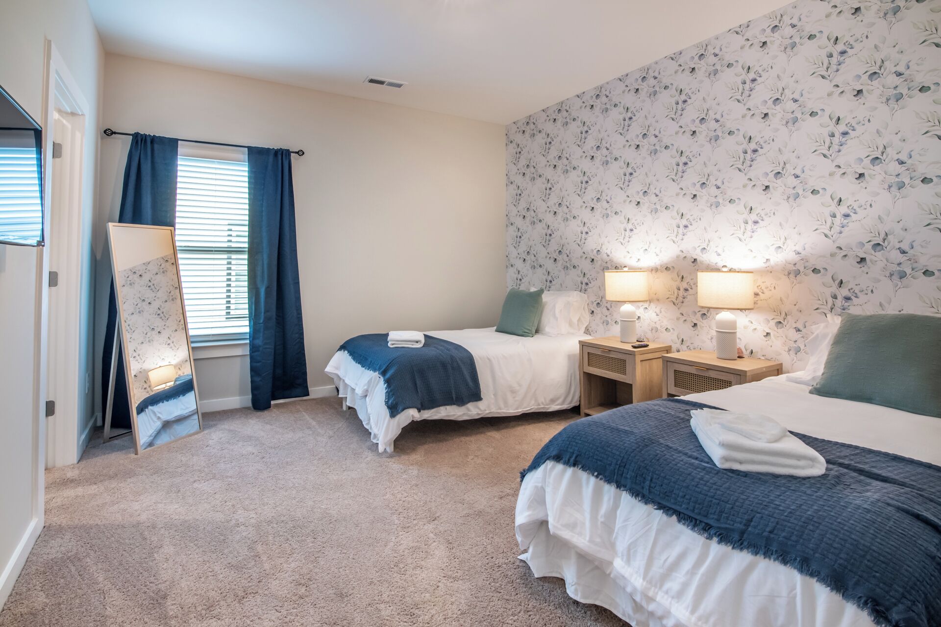 Une chambre d’hôtel avec deux lits, un miroir, une fenêtre avec des rideaux et un papier peint à motifs floraux.
