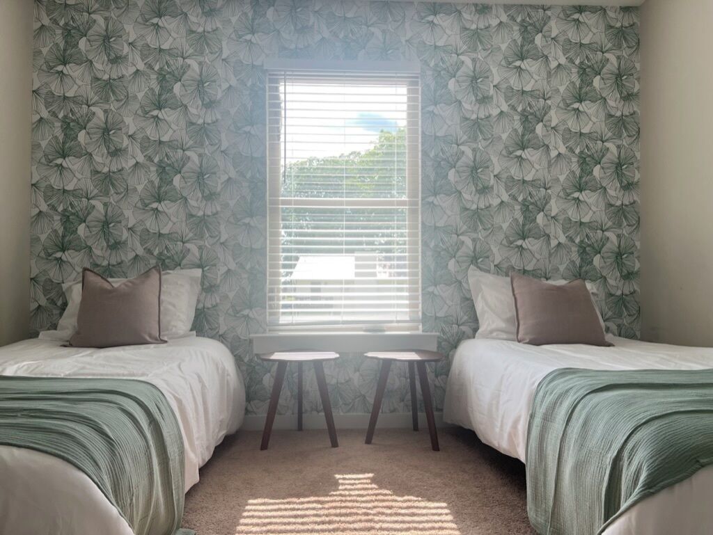 A bedroom with two beds, a window with blinds, and a small table between the beds.