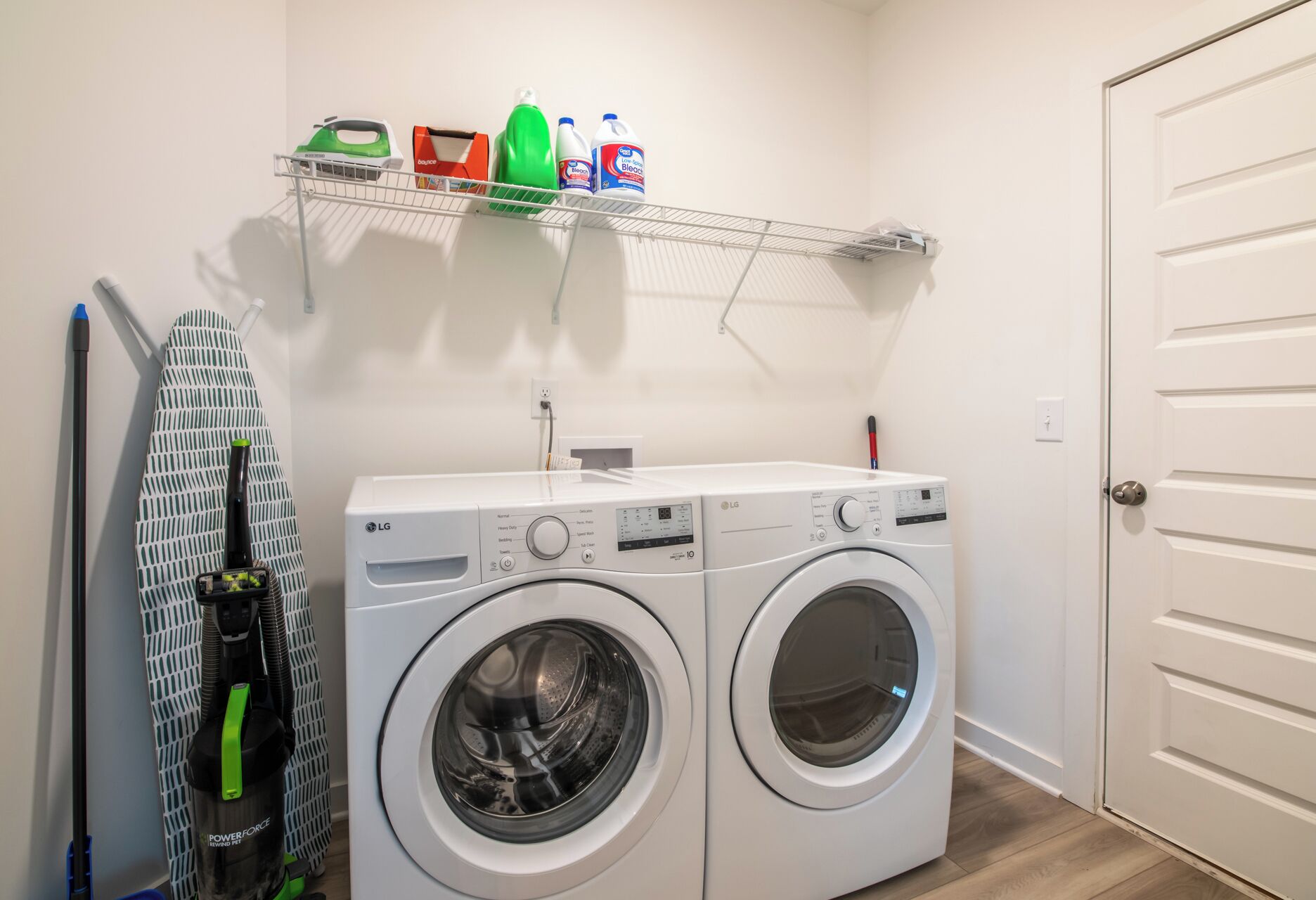 A laundry room with two LG washing machines, a shelf with cleaning supplies, and a hanging rack with ironing boards.