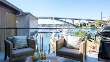 A balcony with two cushioned chairs, a wooden table with books and bottles, and a view of a marina with boats and a bridge.