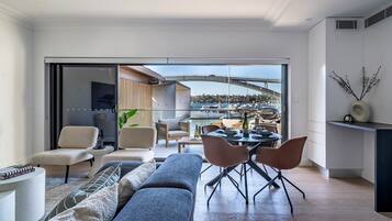 A modern living room with a sofa, chairs, and a dining table. There is a view of a marina with boats and a bridge in the background.