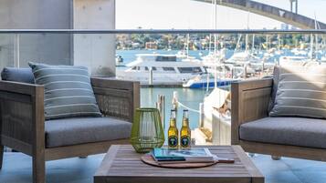 A balcony with two sofas, a table, and bottles of beer, overlooking a marina with boats and a bridge.