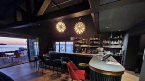 A bar area with a wooden floor, bar stools, and a bar counter. There are hanging lights above the bar and a view of the outdoors through large windows. - La Villa Mandragore (Hyères)