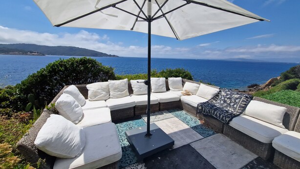 A patio with a white umbrella, wicker sofa, and a view of the sea. - La Villa Mandragore (Hyères)