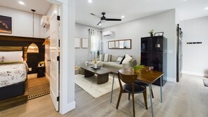 A modern living room with a sofa, coffee table, and dining table. There is a bed with a headboard and a ceiling fan.