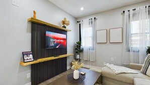 A modern living room with a flat-screen TV, a wooden coffee table, a beige sofa, and a large window with sheer curtains.