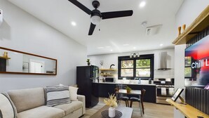 A modern living room with a sofa, coffee table, and a dining area with chairs. A kitchen with black cabinets and a window is visible in the background.