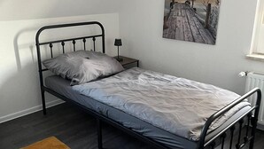 En enkelt seng med metalramme, et brunt gulvtæppe, et natbord med en lampe og et maleri af en solnedgang på væggen.