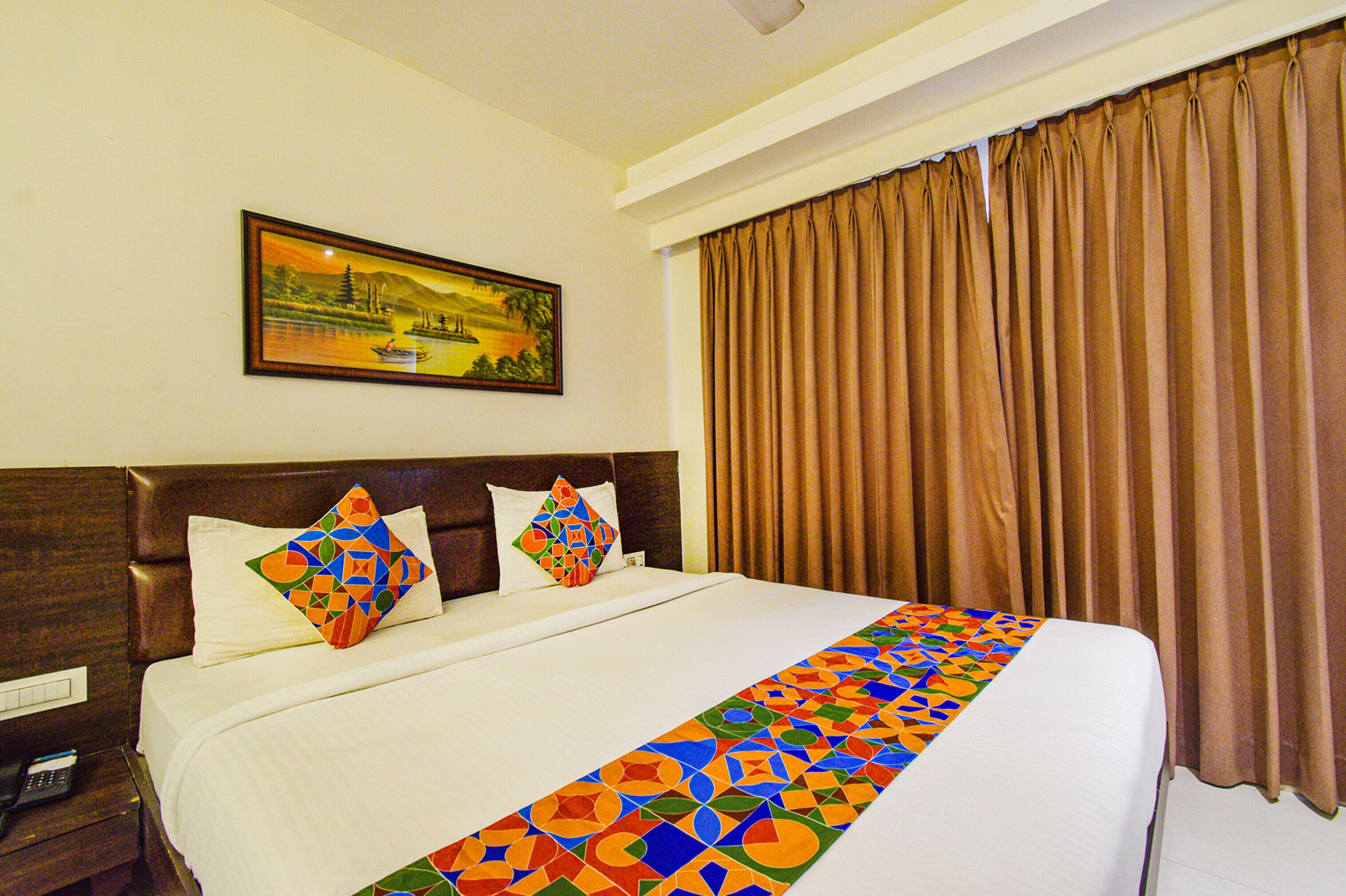 A hotel room with a large bed, two pillows, a painting on the wall, and brown curtains.