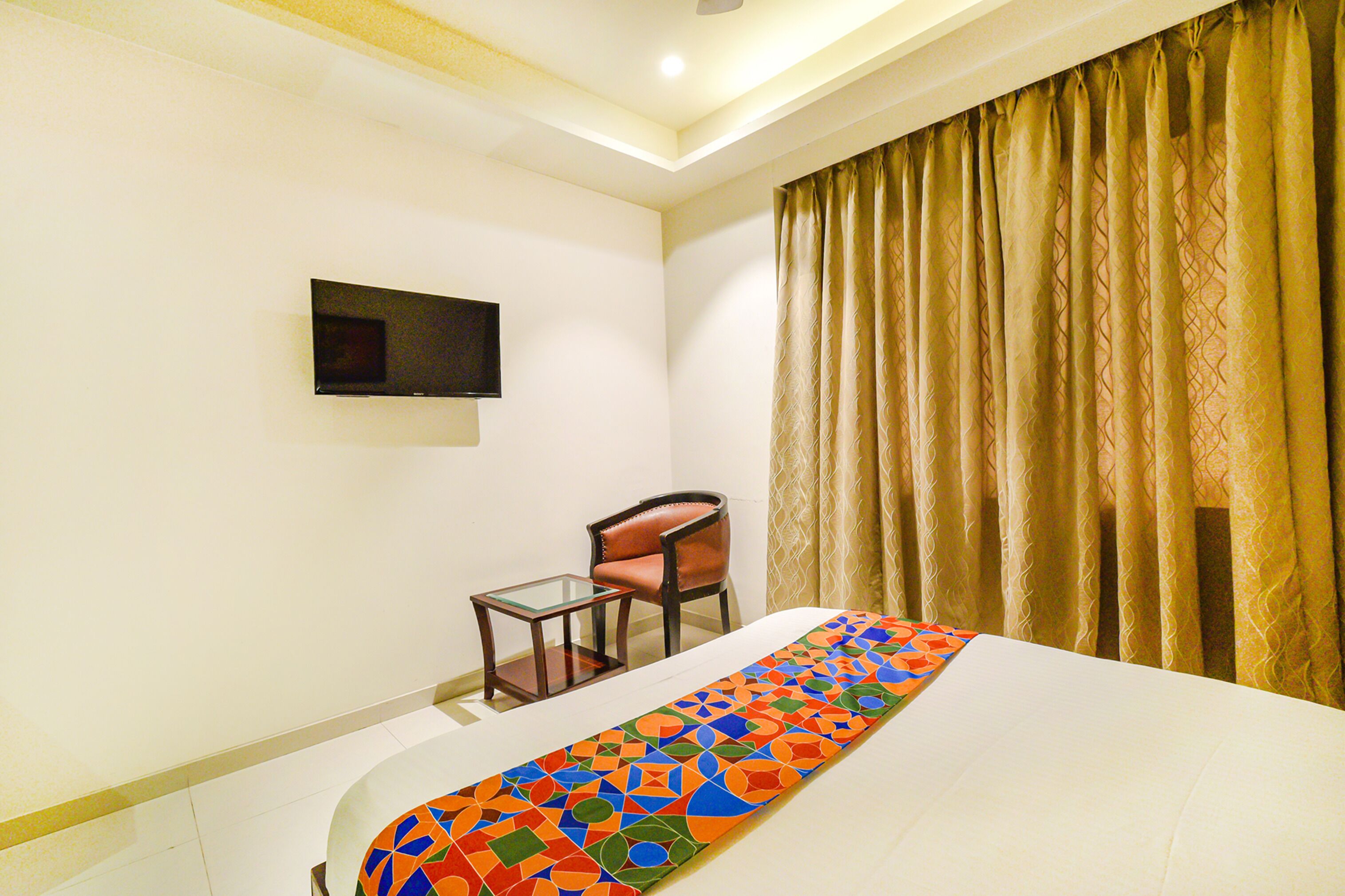 A hotel room with a bed, a small table, a chair, a television mounted on the wall, and a window with curtains.