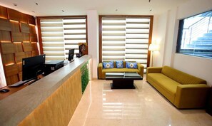 A reception area with a wooden reception desk, a sofa, and a window with blinds. - FabHotel Prime Bellmont (Noida)