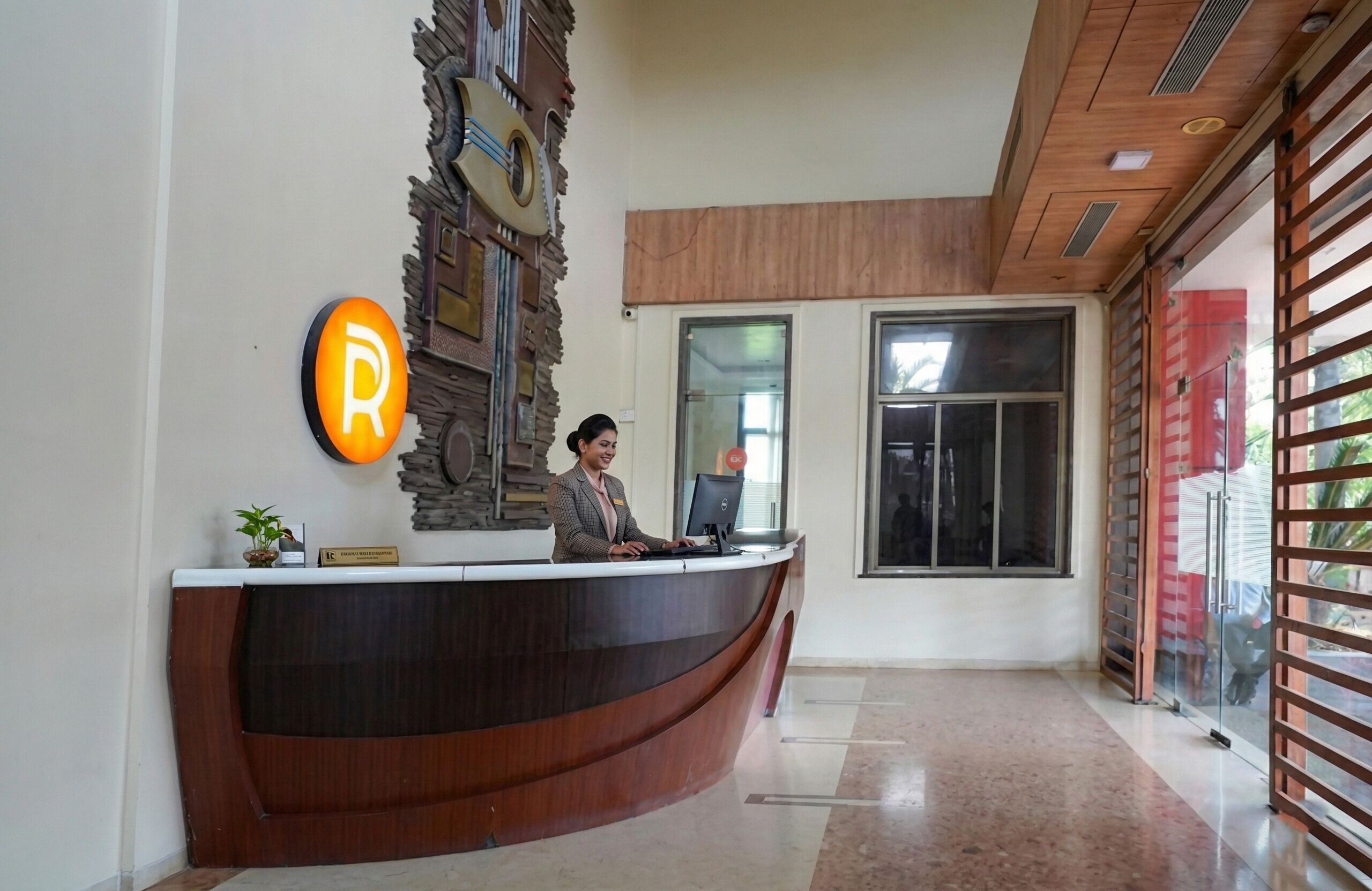A reception desk with a receptionist, a 'R' sign, a decorative wall, and a large window.