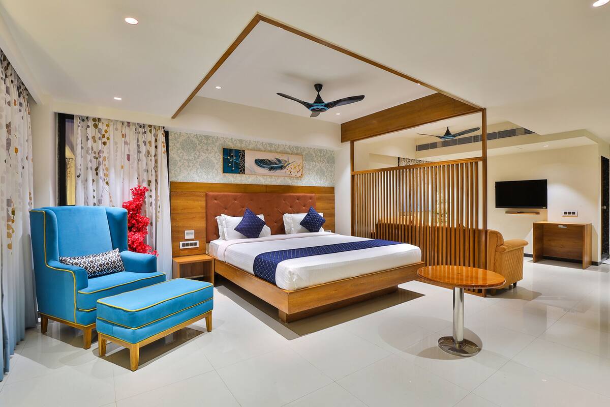 A modern hotel room with a large bed, a blue armchair, a wooden headboard, and a ceiling fan.