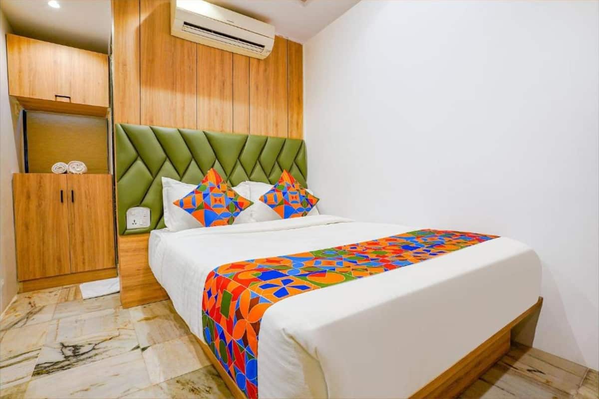 A bedroom with a green upholstered headboard, a bed with white bedding and colorful geometric-patterned throw, a wooden wardrobe, and a marble-patterned floor.