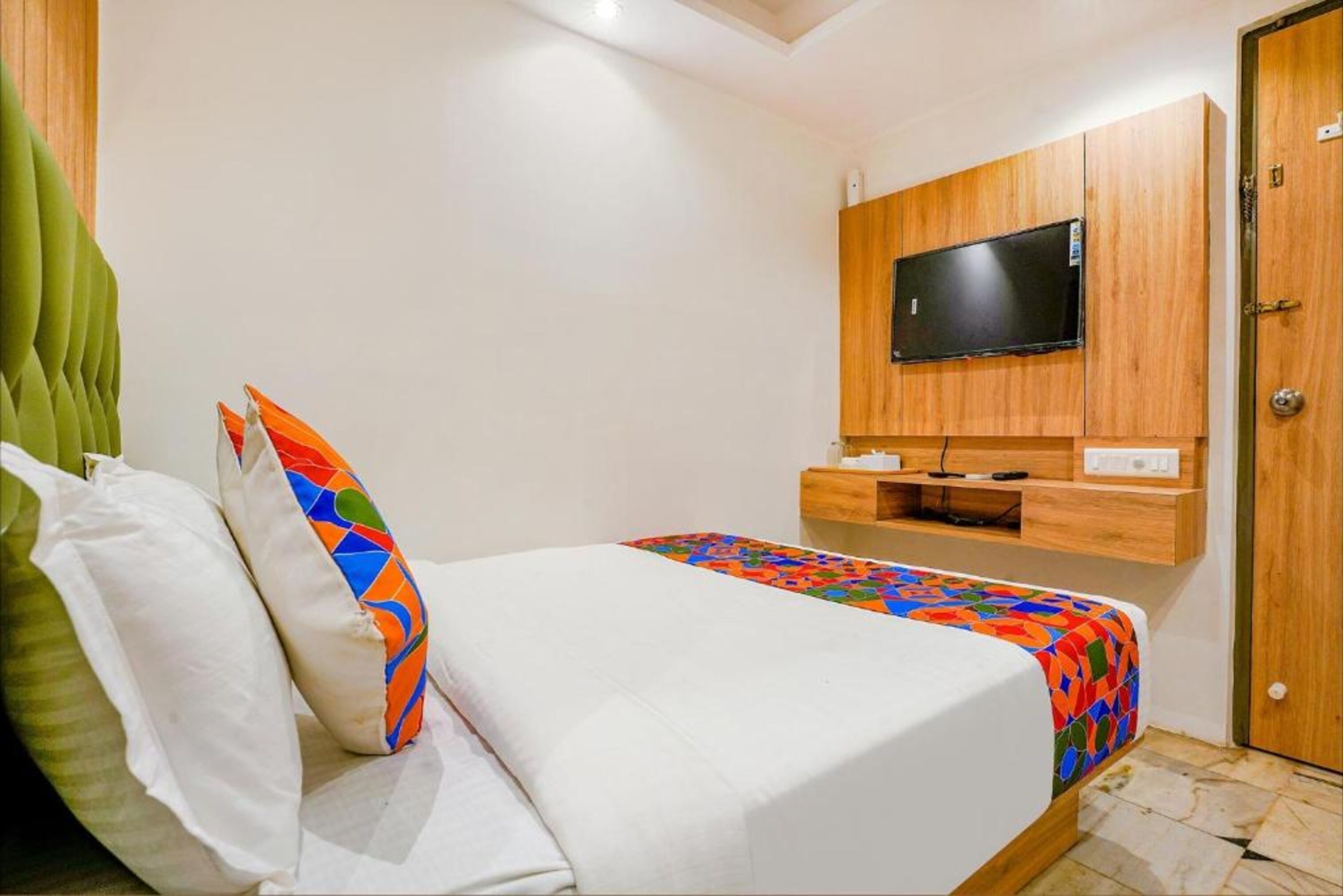 A neatly made bed with colorful bedding, a television mounted on a wooden cabinet, and a green upholstered headboard.