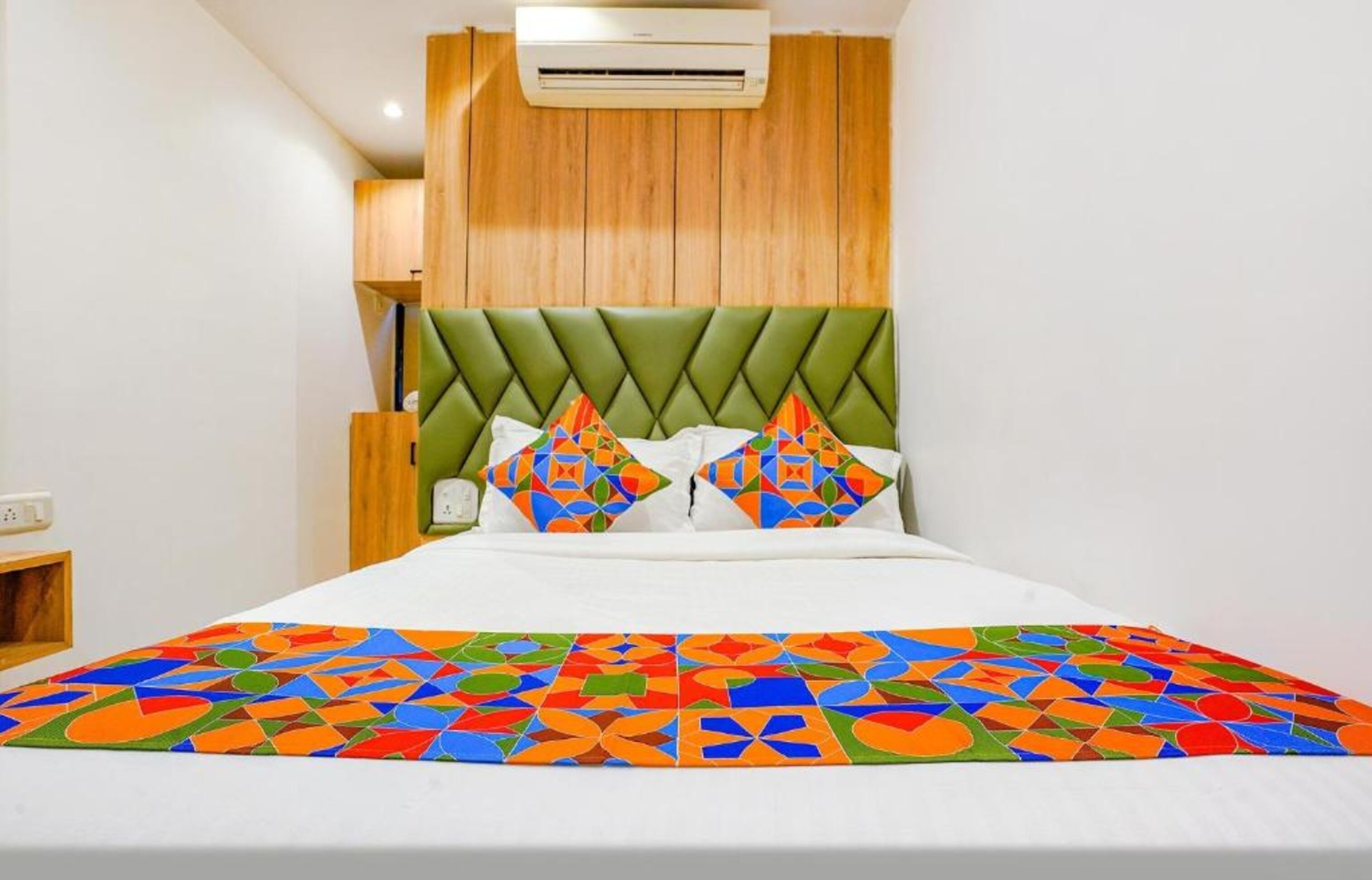 A hotel room with a green upholstered headboard, a colorful geometric-patterned bedspread, and a white pillow with a geometric design.