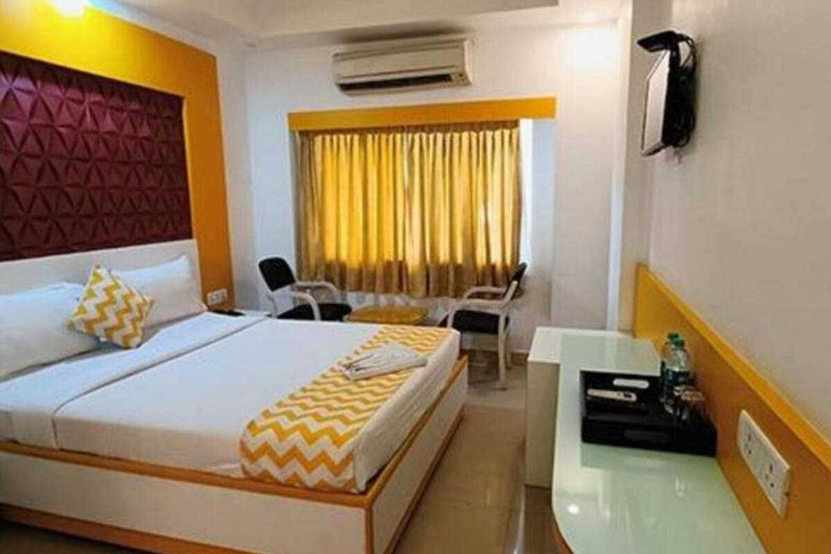 A hotel room with a bed, a desk, a chair, a window with curtains, and an air conditioning unit.