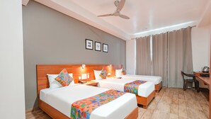 Quarto de hotel com duas camas, uma escrivaninha, uma cadeira, um ventilador de teto e uma janela com cortinas.