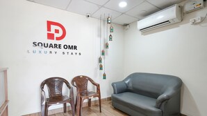 Quarto com sofá cinza, duas cadeiras de madeira, ar condicionado fixado na parede e uma placa com a inscrição 'SQUARE OMR LUXURY STAYS'.