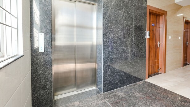 Elevador moderno com acabamento metálico, próximo a uma porta de madeira, em um corredor com piso de pedra polida e parede em tom claro.