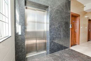 A modern elevator with a metallic finish, adjacent to a wooden door, in a hallway with polished stone flooring and a light-colored wall. - FabHotel D Square OMR (Tirupporur)