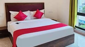 A hotel room with a large bed, a wooden headboard, and a view of buildings through a window with yellow curtains. - FabHotel KSP Kings Inn (Bengaluru)