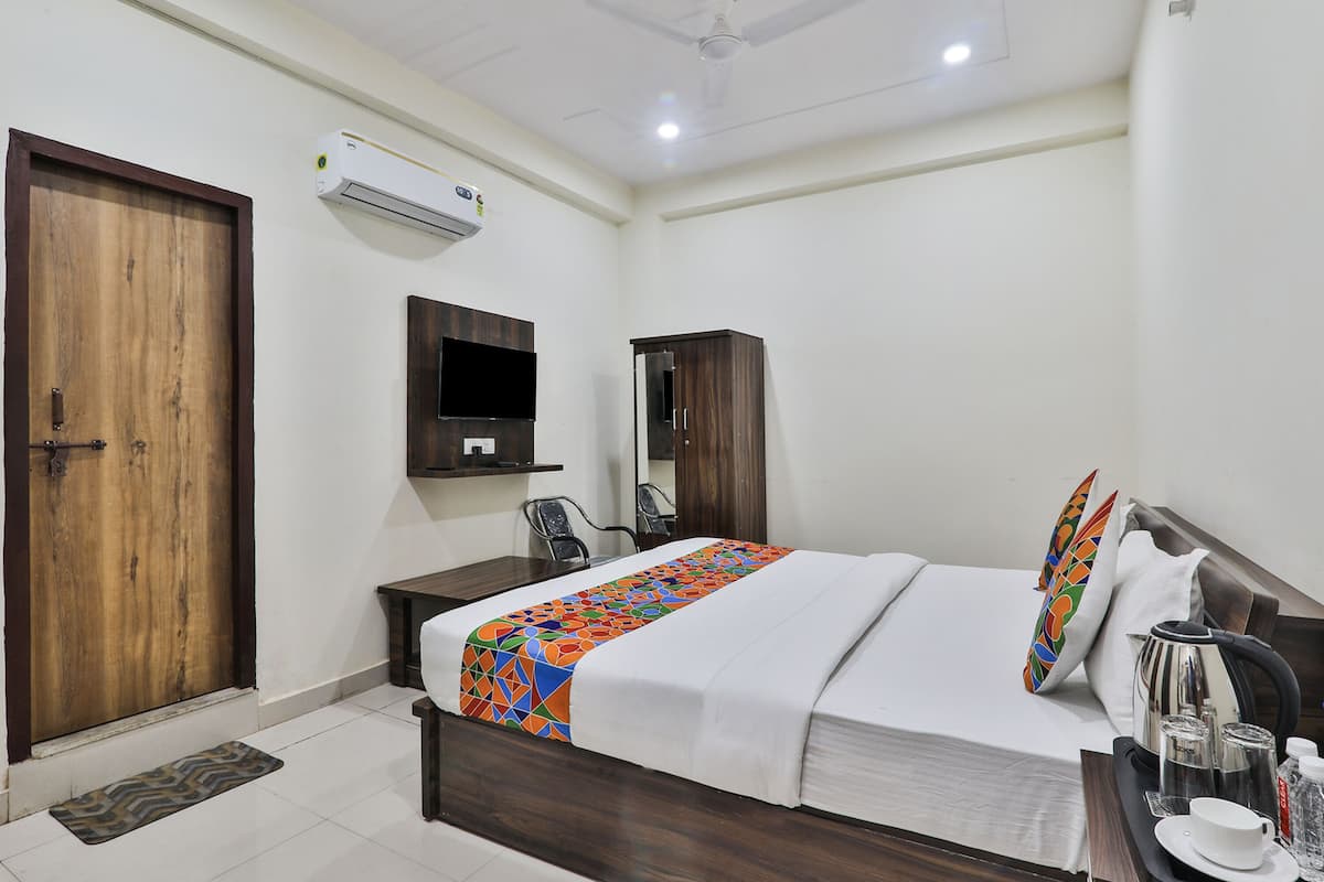 Habitación de hotel con puerta de madera, cama con colcha colorida, televisor montado en la pared, armario, escritorio con silla y cafetera en la mesita de noche.